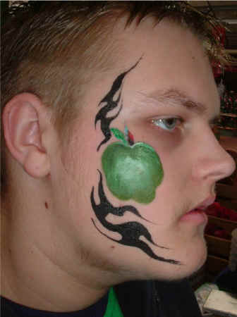 Face Painting: Adult male with cheek painted green apple framed in black trible pattern.