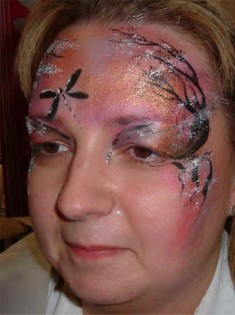 Face Painting: Adult female with pink and peach mask base supporting draggonfly and trees in black with white highlights.
